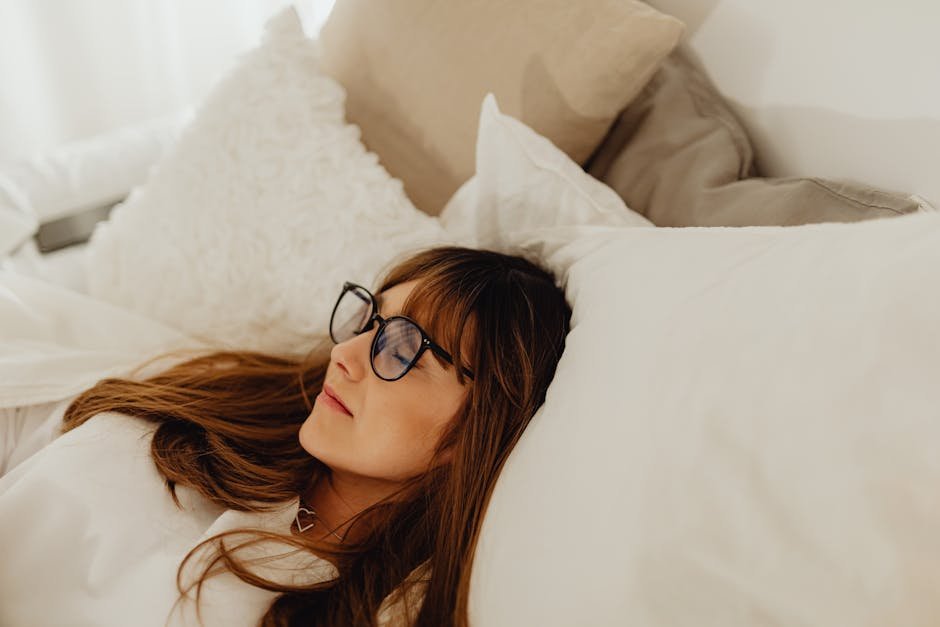 A calm woman wearing eyeglasses lying peacefully in a cozy bed with white pillows.