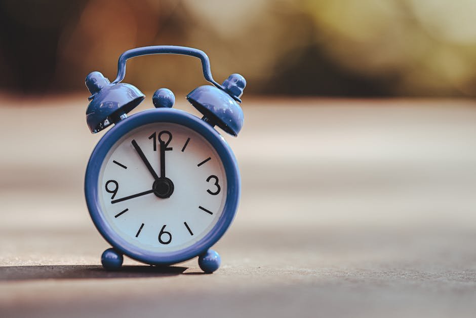 A classic blue alarm clock placed outdoors, displaying five minutes to twelve.