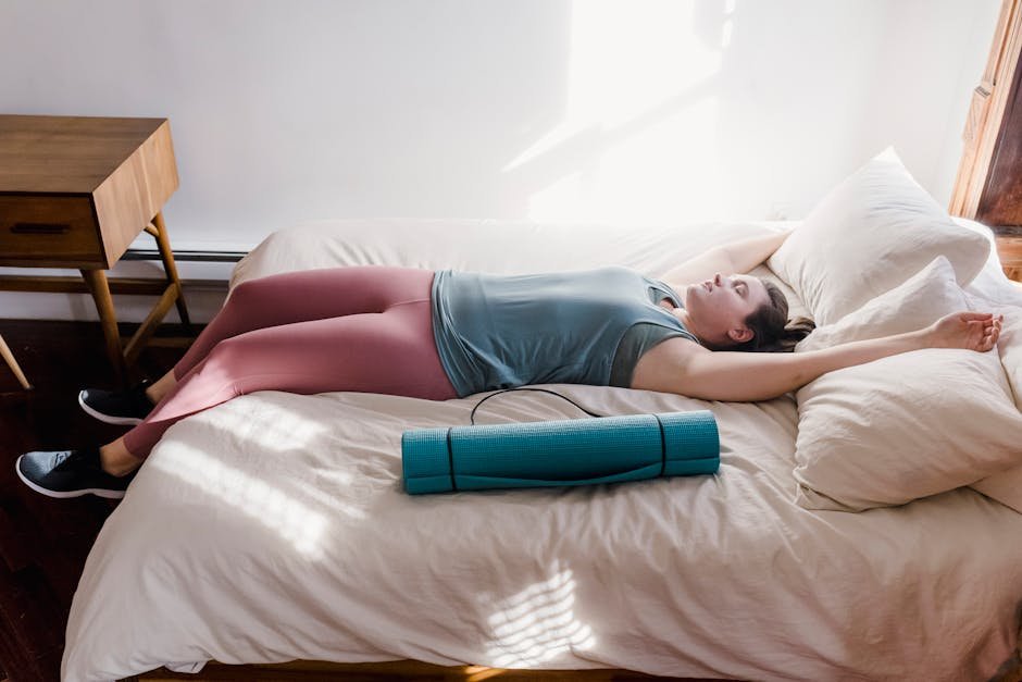 A woman in activewear lying on a bed with a yoga mat, enjoying sunlight and relaxation indoors.