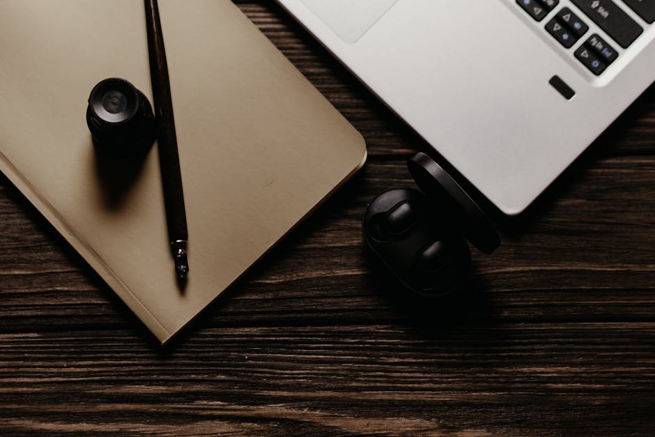 A top-view still life of a laptop, notebook, fountain pen, and earbuds on a wooden table.
