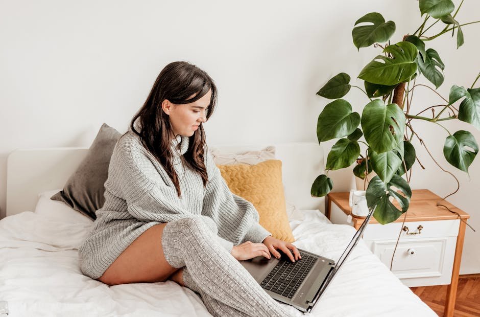 Young woman in cozy sweater, using laptop on bed with plant nearby.