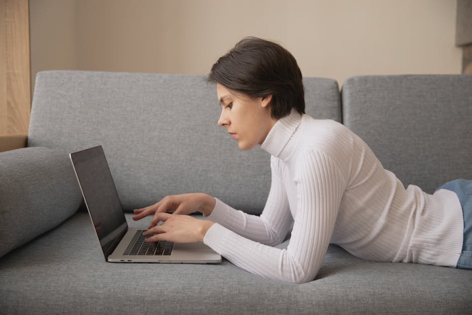 Young adult woman using laptop on a gray sofa indoors, engaged in remote work or leisure activities.