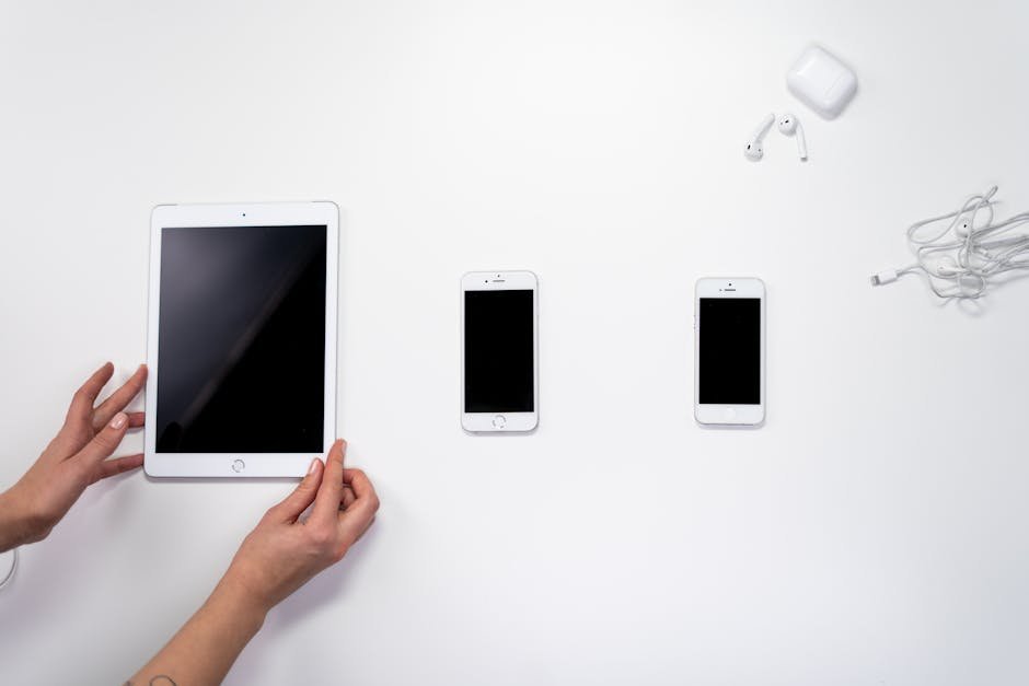 Top view of white digital devices including a tablet, smartphones, and earphones on a white surface.