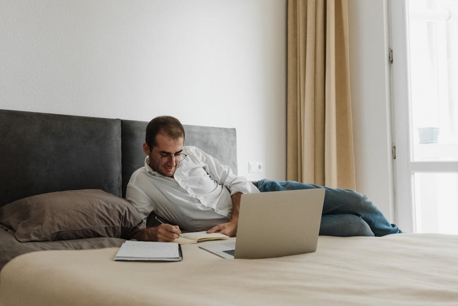 Adult man working remotely on laptop from bed, surrounded by notes and documents.