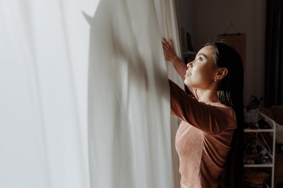 A woman embraces daylight near a white curtain, creating a serene indoor atmosphere.