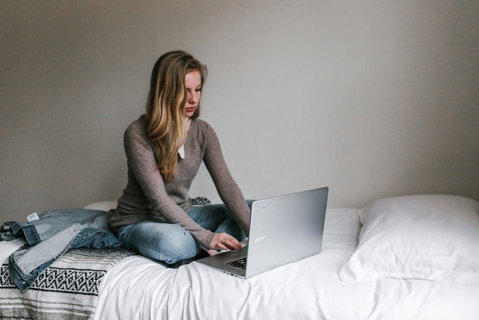A young woman working remotely on a laptop while sitting on a bed in a cozy, modern bedroom.