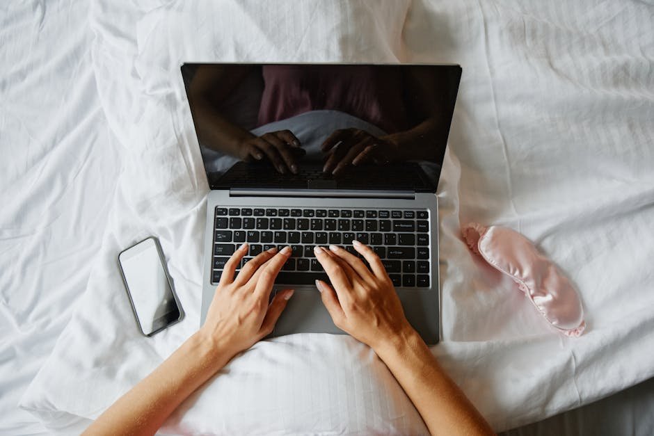 Person lying in bed using a laptop, with a phone and sleep mask nearby, creating a cozy indoor work environment.