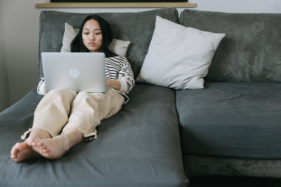 Young Asian woman working remotely on a couch using a laptop, embodying relaxed home office vibes.