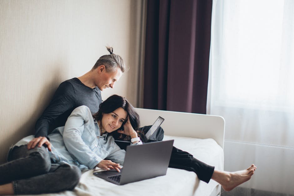 Happy couple working remotely on laptops in a cozy bedroom setting.